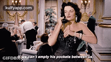 A Woman In A Black Dress Is Talking On A Phone In A Room . GIF