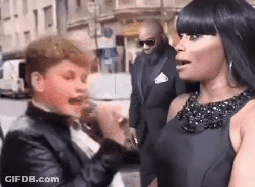 A Woman In A Black Dress Is Talking To A Boy In A Tuxedo On The Street . GIF