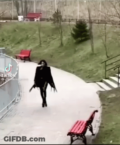A Woman In A Black Dress Is Walking Down A Path With A Red Bench In The Background . GIF