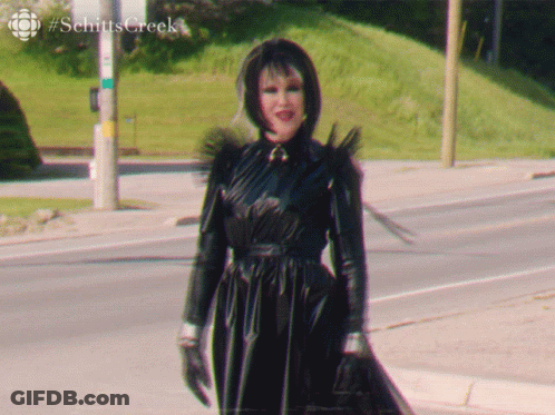 A Woman In A Black Dress Is Walking Down A Street With #Schitts Creek Written In The Corner GIF