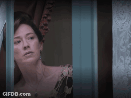 A Woman In A Black Dress Looks Out A Window With A Blue Curtain Behind Her GIF