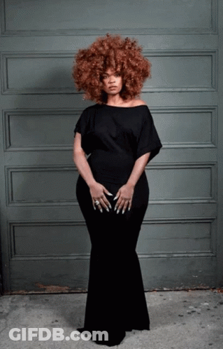 A Woman In A Black Dress Stands In Front Of A Gray Wall GIF
