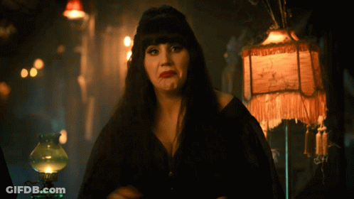 A Woman In A Black Dress Stands In Front Of A Lamp With A Fringed Shade That Says ' A ' GIF