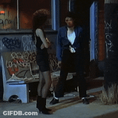 A Woman In A Black Dress Stands Next To A Man In A Blue Jacket In Front Of A Building With Graffiti On It GIF