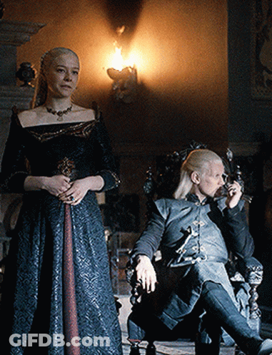 A Woman In A Black Dress Stands Next To A Man In A Chair Smoking A Cigarette GIF