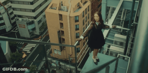 A Woman In A Black Dress Stands On A Balcony Overlooking A City With A Building That Says Comic Shop On It GIF