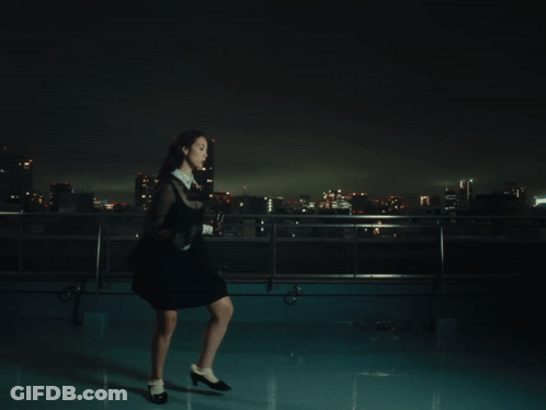 A Woman In A Black Dress Stands On A Rooftop At Night GIF