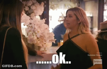 A Woman In A Black Dress Talks To Another Woman In Front Of A Sign That Says Ok On It GIF