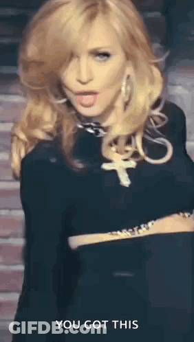 A Woman In A Black Dress With A Cross Necklace Is Standing In Front Of A Brick Wall And Says You Got This . GIF