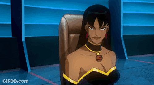 A Woman In A Black Dress With A Gold Necklace With The Letter S On It Sits In A Chair GIF