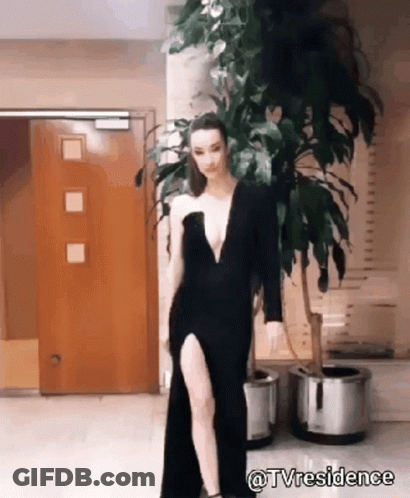 A Woman In A Black Dress With A High Slit Is Standing In Front Of A Plant . GIF