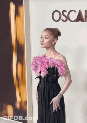 A Woman In A Black Dress With Pink Feathers Stands In Front Of A Sign That Says Oscar GIF