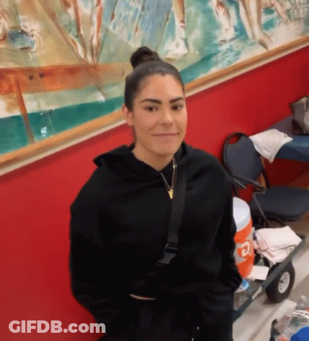 A Woman In A Black Hoodie Is Standing In Front Of A Red Wall And Smiling . GIF