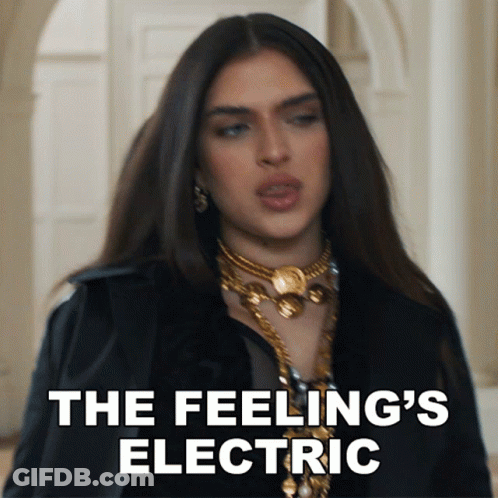 A Woman In A Black Jacket And Gold Necklace Says " The Feeling 'S Electric " In White Letters GIF
