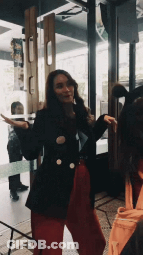 A Woman In A Black Jacket And Red Pants Is Standing In Front Of A Door GIF