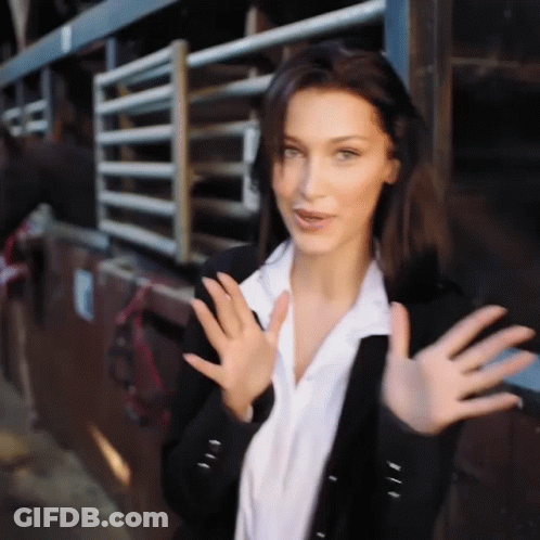 A Woman In A Black Jacket And White Shirt Is Waving Her Hands In Front Of A Horse Stable . GIF