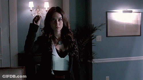 A Woman In A Black Jacket And White Top Stands In A Dark Room GIF