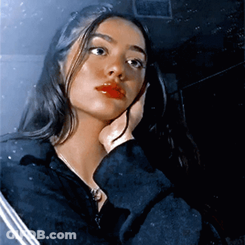 A Woman In A Black Jacket Holds Her Hand To Her Face And Looks At The Camera GIF