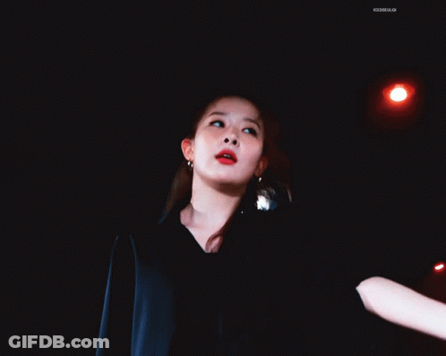A Woman In A Black Jacket Is Dancing In A Dark Room With A Blue Light Behind Her GIF