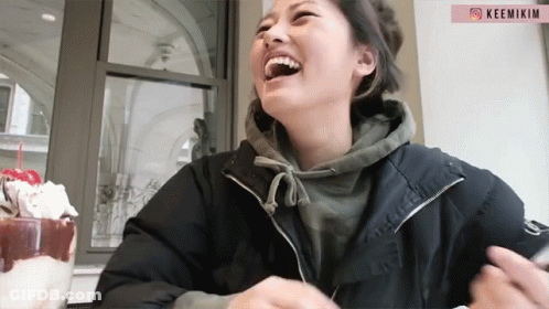 A Woman In A Black Jacket Is Laughing In Front Of A Pink Sign That Says Keemikm GIF