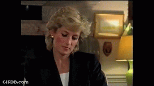 A Woman In A Black Jacket Is Sitting At A Table With A Lamp In The Background . GIF