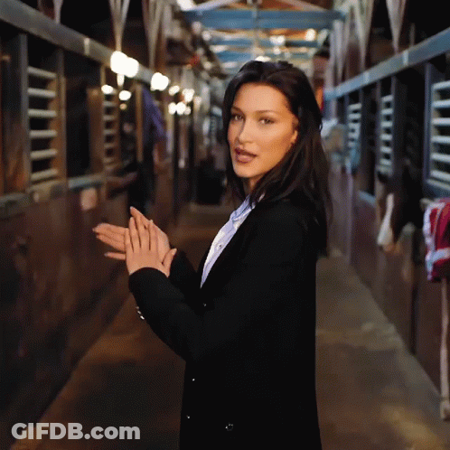 A Woman In A Black Jacket Is Standing In A Stable With Her Hands Folded GIF