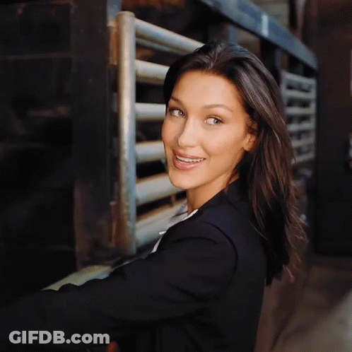 A Woman In A Black Jacket Smiles In Front Of A Metal Fence GIF