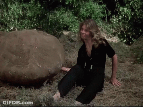 A Woman In A Black Jumpsuit Sits In The Grass Next To A Large Rock GIF