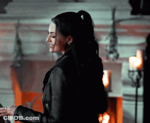 A Woman In A Black Leather Jacket Is Smiling In Front Of Candles GIF
