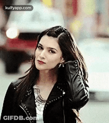 A Woman In A Black Leather Jacket Is Standing On A Street Holding Her Hair . GIF