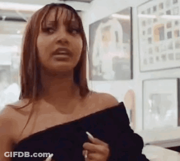 A Woman In A Black Off The Shoulder Top Is Standing In Front Of A Wall With Paintings On It . GIF