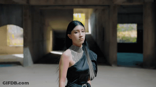 A Woman In A Black One Shoulder Dress Stands In A Dark Room GIF