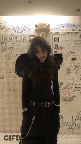 A Woman In A Black Outfit Is Standing In Front Of A Wall That Has Graffiti On It Including The Word Ovoks GIF