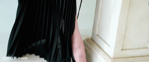 A Woman In A Black Pleated Skirt Is Standing Next To A White Column GIF