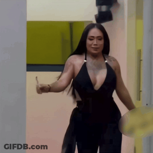A Woman In A Black Plunging Neckline Top Is Standing In A Hallway GIF