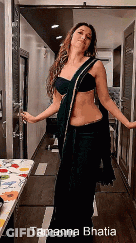 A Woman In A Black Saree Is Standing In A Hallway With The Name Tamanna Bhatia On The Bottom Right GIF