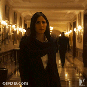 A Woman In A Black Scarf Is Walking Down A Hallway With A Man In A Suit Behind Her GIF