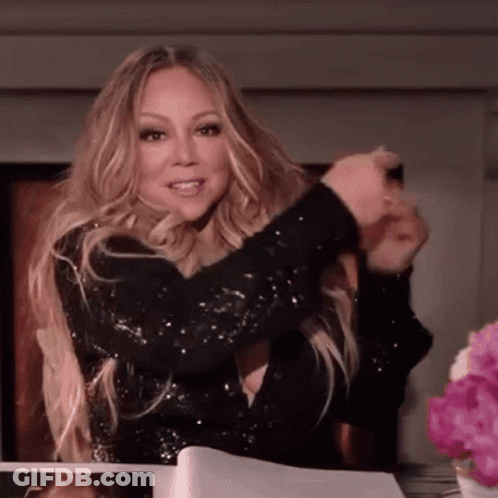 A Woman In A Black Sequined Dress Is Sitting At A Table With Flowers In Front Of Her GIF