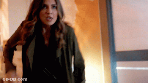 A Woman In A Black Shirt And A Green Jacket Is Standing In A Hallway . GIF