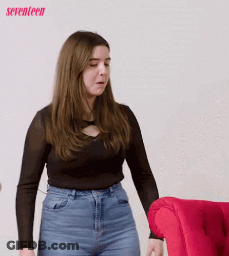 A Woman In A Black Shirt And Blue Jeans Is Standing In Front Of A Red Couch . GIF