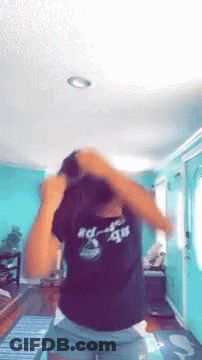 A Woman In A Black Shirt And Shorts Is Standing In A Living Room With Her Hair In A Bun . GIF