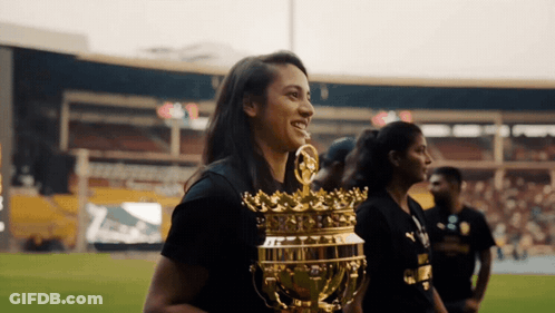 A Woman In A Black Shirt Holds A Gold Trophy With A Crown On It GIF