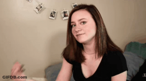 A Woman In A Black Shirt Is Sitting On A Bed In Front Of A Wall With Pictures Hanging On It . GIF
