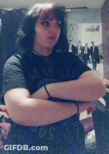 A Woman In A Black Shirt Is Standing With Her Arms Crossed In Front Of A Beatles Poster . GIF