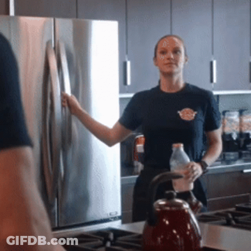 A Woman In A Black Shirt Stands In A Kitchen Holding A Bottle GIF