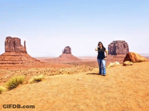 A Woman In A Black Shirt Stands On A Dirt Road In The Desert GIF