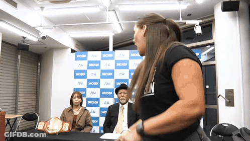A Woman In A Black Shirt That Says ' N ' On It Stands In Front Of A Blue And White Backdrop GIF