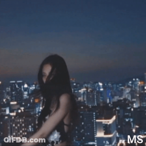 A Woman In A Black Strapless Dress Is Standing In Front Of A City Skyline At Night . GIF
