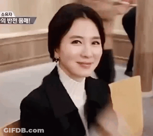 A Woman In A Black Suit And White Shirt Is Sitting In A Chair And Smiling . GIF