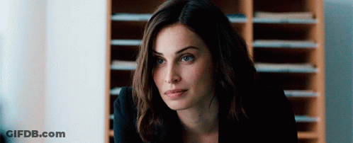 A Woman In A Black Suit Is Sitting At A Desk In Front Of A Bookshelf And Looking At The Camera . GIF